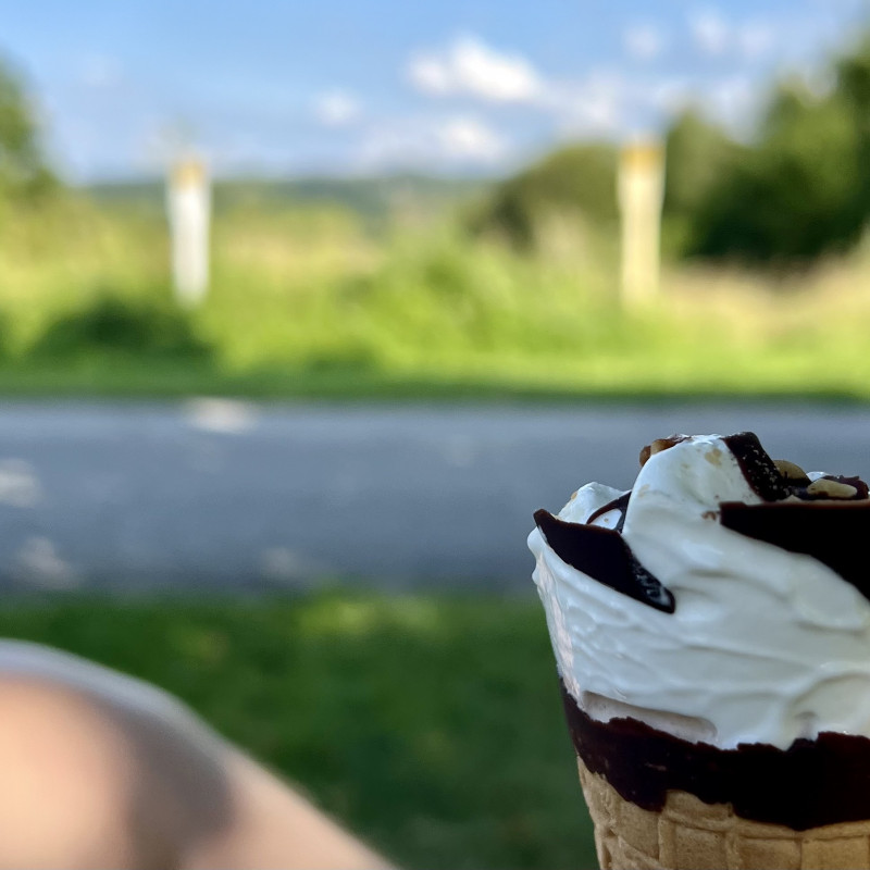 Vanilleeis mit Schokoladenüberzug in einer Waffel, ein Knie und im Hintergrund der Blick über grüne Wiesen bei blauem Himmel.