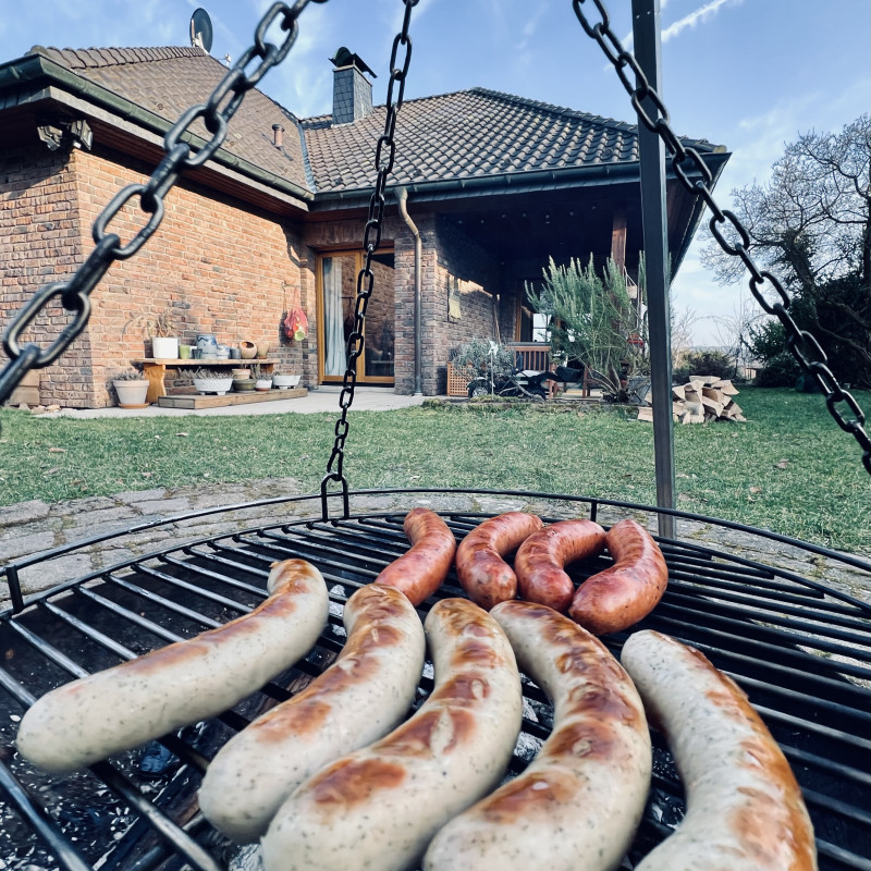 Grillwürste liegen auf einem Rost über Glut, im Hintergrund eine grüne Wiese und ein verklinkertes Haus vor blauem Himmel.